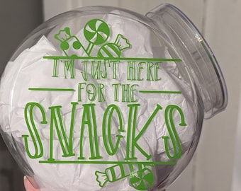 May include: A clear, round candy jar with a lid, filled with white tissue paper. The jar has green text that reads "I'M JUST HERE FOR THE SNACKS" with candy graphics.