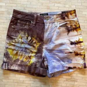 May include: A pair of denim shorts with a tie-dye pattern. The shorts are split down the middle, with one side being a brown and yellow tie-dye and the other side being a blue and white tie-dye. The shorts have a cuffed hem and a high waist.