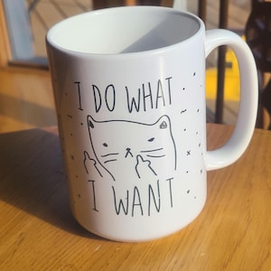 May include: White ceramic mug with a black cat illustration and the text "I DO WHAT I WANT." The cat is depicted with its middle fingers up. The mug has a large handle and is sitting on a wooden surface.