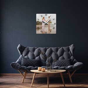 May include: Square wall clock featuring a beach scene with a couple holding hands. The clock has black Roman numerals and black clock hands. The clock is mounted on a dark blue wall above a gray sofa and wooden table.