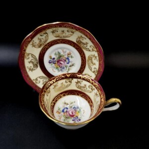 May include: A vintage teacup and saucer set. The cup and saucer are cream-coloured with a maroon border and gold accents. Floral designs are featured in the centre of the cup and saucer. The cup has a gold handle.