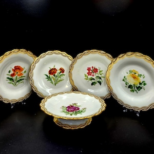 May include: A set of five white porcelain plates and a serving dish, each decorated with a unique floral design. The plates have scalloped edges with gold trim. The flowers are painted in shades of red, orange, yellow, and purple.