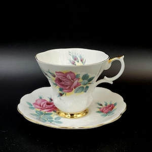 May include: A white teacup and saucer set with a pink rose floral pattern. The cup has a gold rim and the saucer has a scalloped edge.
