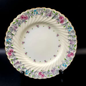 May include: Cream porcelain plate with a gold rim and a scalloped edge. The plate features a floral design with pink roses, purple flowers, and blue accents, surrounded by grey leaves. The plate has a spiral design.