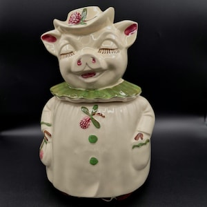 Puede incluir: Un tarro de galletas de cerámica vintage con forma de cerdo sonriente que lleva un sombrero y un cuello verde. El cerdo es de color crema con detalles en rojo en las orejas y el hocico. El sombrero presenta un diseño floral. El tarro tiene un cuerpo blanco con botones verdes y un diseño floral.