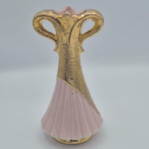 Könnte beinhalten: Eine Keramikvase mit einem blassrosa geriffelten Sockel und einem metallisch goldenen Oberteil mit zwei symmetrischen Henkeln. Der goldene Bereich zeigt ein kompliziertes geprägtes Blumenmuster, während die Basis glatt und gerippt ist.