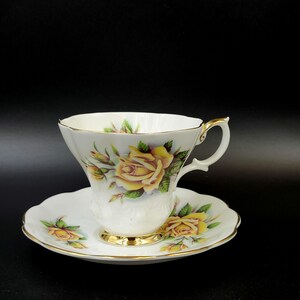 May include: A white teacup and saucer with a gold rim. The cup and saucer feature a yellow rose floral pattern.