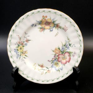 May include: A white ceramic plate with a green and gold rim. The plate features a floral design with pink, yellow, blue, and orange flowers.