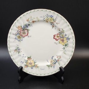 May include: A white ceramic dinner plate with a green and gold rim. The plate features a floral design with colorful flowers and green leaves.