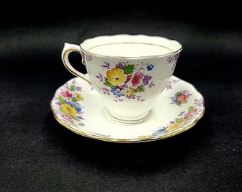 Puede incluir: Una taza de té y un platillo blancos con un patrón floral en tonos de morado, amarillo y rosa. La taza y el platillo tienen un borde dorado.