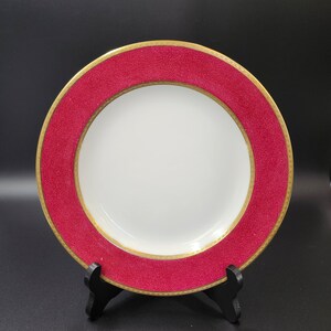 May include: A white plate with a wide, textured, red rim and a gold border. The plate is displayed on a black stand against a black background. The plate is a piece of dinnerware.