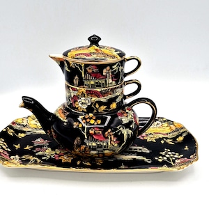 May include: A black ceramic tea set comprising a teapot, two cups, and a rectangular tray. The set showcases a detailed landscape design in gold, red, and yellow. The teapot has a curved spout and handle, and the tray has a gold trim.