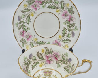 Könnte beinhalten: Ein weißes Porzellan-Tassenset mit Untertasse, gewelltem Rand und Goldverzierung. Das Set ist mit einem Blumenmuster aus rosa Rosen, grünen Blättern und violetten Disteln verziert. Im Inneren der Tasse befinden sich eine goldene Krone und die Buchstaben E R.