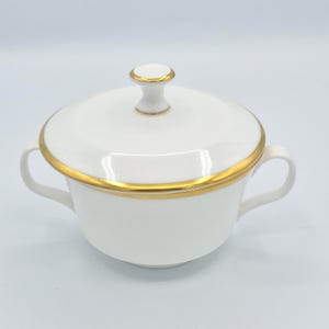 May include: A white porcelain sugar bowl with a matching lid, featuring a smooth finish and a gold-trimmed rim. The bowl includes two curved side handles and a small, round, gold-accented knob on the centre of the lid for easy removal.