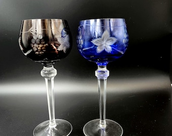 Puede incluir: Dos copas de vino de cristal grabadas, una con un tono morado oscuro y la otra con un tono azul brillante. Ambas copas presentan un diseño de vid blanca.