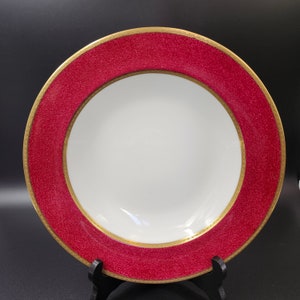 May include: A white plate with a textured red border and a gold rim. The plate is round and sits on a black stand. The plate is designed for serving food.