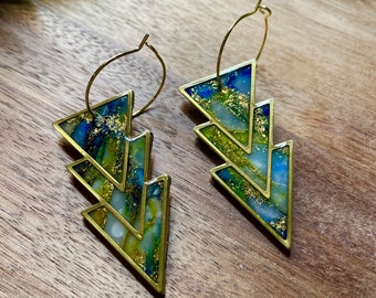 May include: A pair of gold-toned hoop earrings with a layered lightning bolt design. The earrings feature a mix of blue, green, and gold resin, creating a marbled effect. The earrings are set against a wooden background.