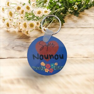 Peut inclure: Un porte-clés rond bleu avec un cœur rouge et le texte "Super Nounou" en noir. Le cœur a le mot "Super" écrit à l'intérieur en blanc. Le porte-clés a un motif floral autour du bas.