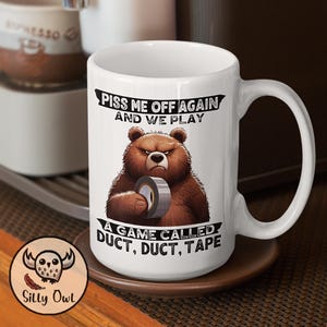 A 15oz white ceramic coffee mug featuring a cartoon-style illustration of a grumpy grizzly bear. The mug displays the bold, humorous text: "Piss Me Off Again And We Play A Game Called Duct Duct Tape." The design is printed on both sides, making it perfect for right- or left-handed users. A fun and sarcastic gift for coffee lovers with a wild sense of humor.