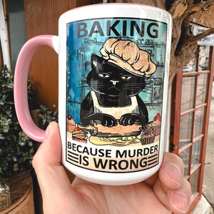 May include: White ceramic mug with a pink handle. The mug features a black cat wearing a chef's hat and apron, rolling out dough. The text on the mug reads "Baking because murder is wrong."