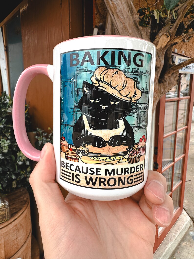 May include: White ceramic mug with a pink handle. The mug features a black cat wearing a chef's hat and apron, rolling out dough. The text on the mug reads "Baking because murder is wrong."
