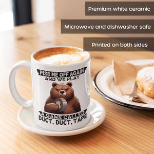 A white ceramic coffee mug featuring a cartoon-style illustration of a grumpy grizzly bear. The mug displays the bold, humorous text: "Piss Me Off Again And We Play A Game Called Duct Duct Tape." The design is printed on both sides, making it perfect for right- or left-handed users. A fun and sarcastic gift for coffee lovers with a wild sense of humor.