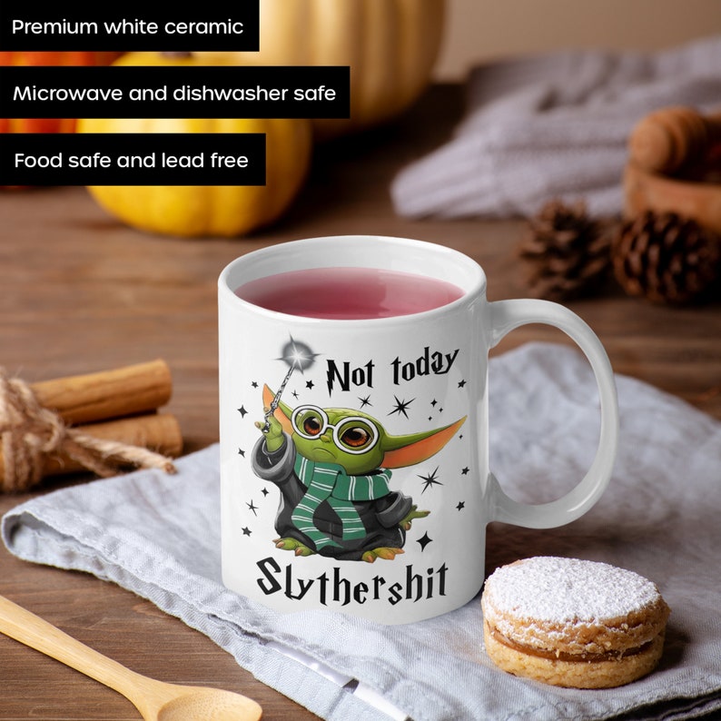 May include: White ceramic mug with a green character wearing glasses and a green and black robe. The character is holding a wand and the mug says "Not today Slythershit".