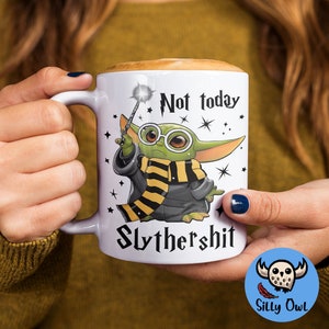 May include: White ceramic mug with a green character wearing glasses and a yellow and black scarf. The character is holding a wand and the mug says "Not today Slythershit".
