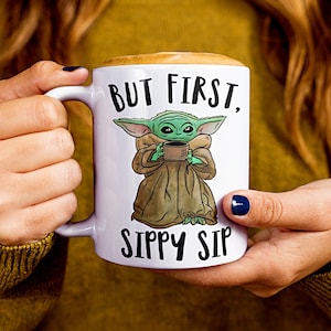 A white ceramic coffee mug featuring a cute illustration of Baby Yoda with the phrase "But First, Sippy Sip." A fun and nerdy mug, perfect for sci-fi fans, coffee lovers, and anyone who needs their caffeine fix before taking on the day.