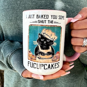 May include: White ceramic mug with a pink handle. The mug features a black cat wearing a chef's hat and baking cupcakes. The text on the mug reads "I just baked you some -SHUT THE- FUCUPCAKES".