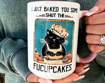 May include: White ceramic mug with a pink handle. The mug features a black cat wearing a chef's hat and baking cupcakes. The text on the mug reads "I just baked you some -SHUT THE- FUCUPCAKES".