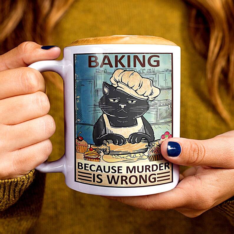 May include: White ceramic mug with a black cat wearing a chef's hat and apron. The mug has a colorful illustration of the cat baking in a kitchen. The text on the mug reads "Baking because murder is wrong."