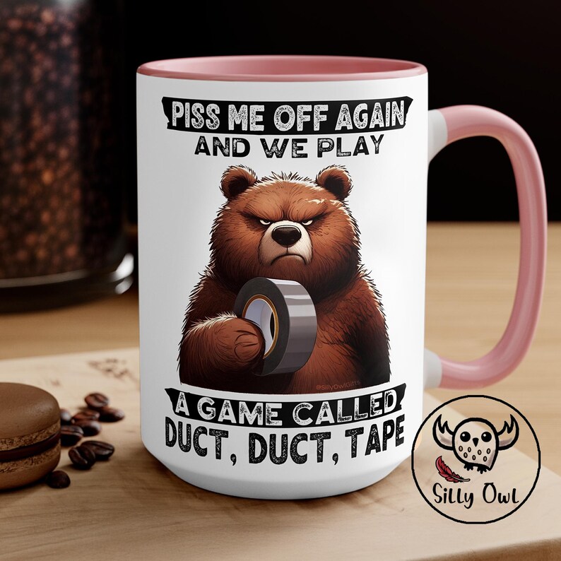 A 15oz pink and white ceramic coffee mug featuring a cartoon-style illustration of a grumpy grizzly bear. The mug displays the bold, humorous text: "Piss Me Off Again And We Play A Game Called Duct Duct Tape." The design is printed on both sides, making it perfect for right- or left-handed users. A fun and sarcastic gift for coffee lovers with a wild sense of humor.