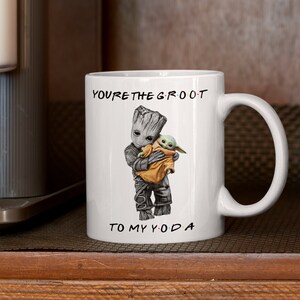 A white ceramic coffee mug featuring an adorable illustration of Groot holding Baby Yoda with the phrase "You're the Groot to My Yoda." A fun and heartwarming sci-fi crossover mug, perfect for best friends, pop culture fans, and geeky coffee lovers.