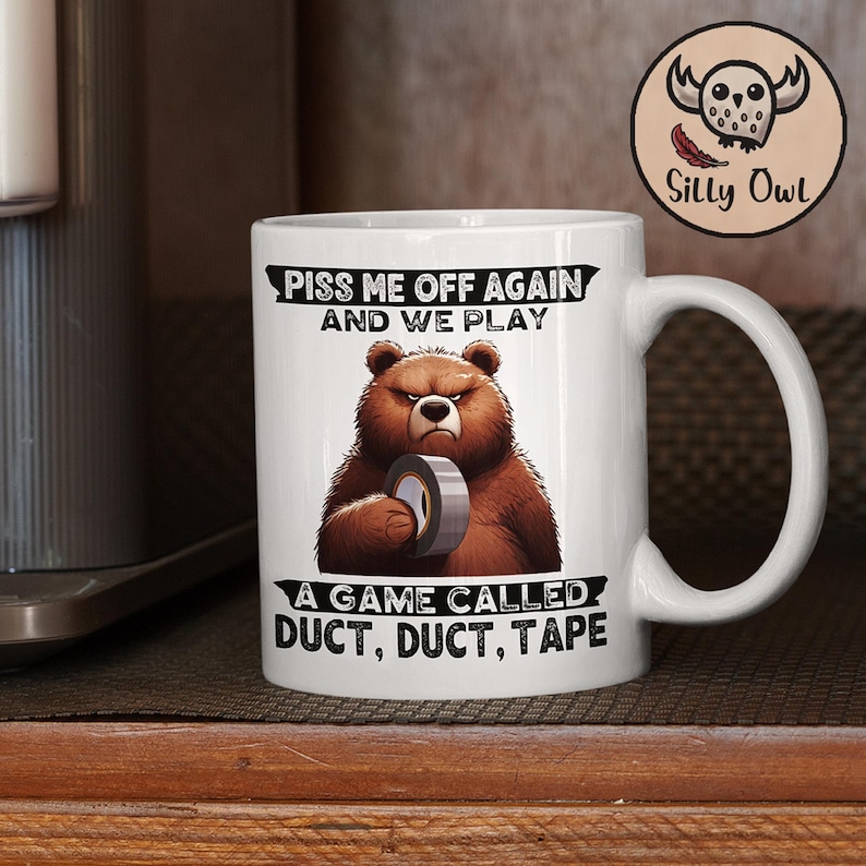 A white ceramic coffee mug featuring a cartoon-style illustration of a grumpy grizzly bear. The mug displays the bold, humorous text: "Piss Me Off Again And We Play A Game Called Duct Duct Tape." The design is printed on both sides, making it perfect for right- or left-handed users. A fun and sarcastic gift for coffee lovers with a wild sense of humor.