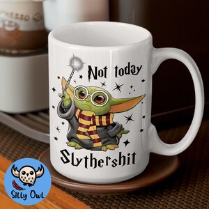 May include: White ceramic mug with a green cartoon character holding a wand and wearing a black robe with a yellow and red scarf. The text "Not today Slythershit" is printed on the mug.