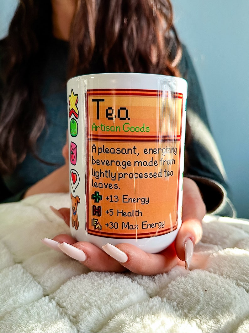 K&ouml;nnte beinhalten: Eine wei&szlig;e Keramiktasse mit einem bunten Design mit dem Text "Tea Artisan Goods". Das Design enth&auml;lt ein Pixelbild eines Teeblattes und eine Liste von Vorteilen: +13 Energy, +5 Health, +30 Max Energy.