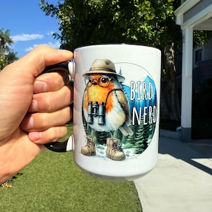 May include: White ceramic mug with a black handle. The mug features a cartoon robin wearing a hat, glasses, and boots. The robin is holding binoculars and is surrounded by trees. The text "Bird Nerd" is printed on the mug.