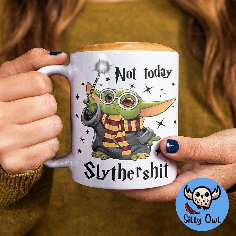 May include: White ceramic mug with a green alien character holding a wand and wearing a Hogwarts house scarf. The mug has the text "Not today Slythershit" printed on it.