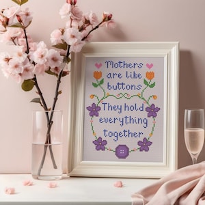 May include: A framed cross-stitch artwork with the text "Mothers are like buttons. They hold everything together." The design features floral and button motifs in purple, orange, and green. A vase of pink flowers and a glass of champagne are in the background.