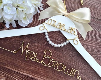 May include: A white wooden clothes hanger featuring a gold wire script reading Mrs. Brown and the date 07.26.25. The hanger is adorned with a cream-colored satin ribbon bow and a circular accent of white faux pearls.