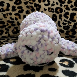 May include: A plush toy shaped like a seal with a white, pink, and purple speckled fur. The toy has black eyes and a closed mouth.