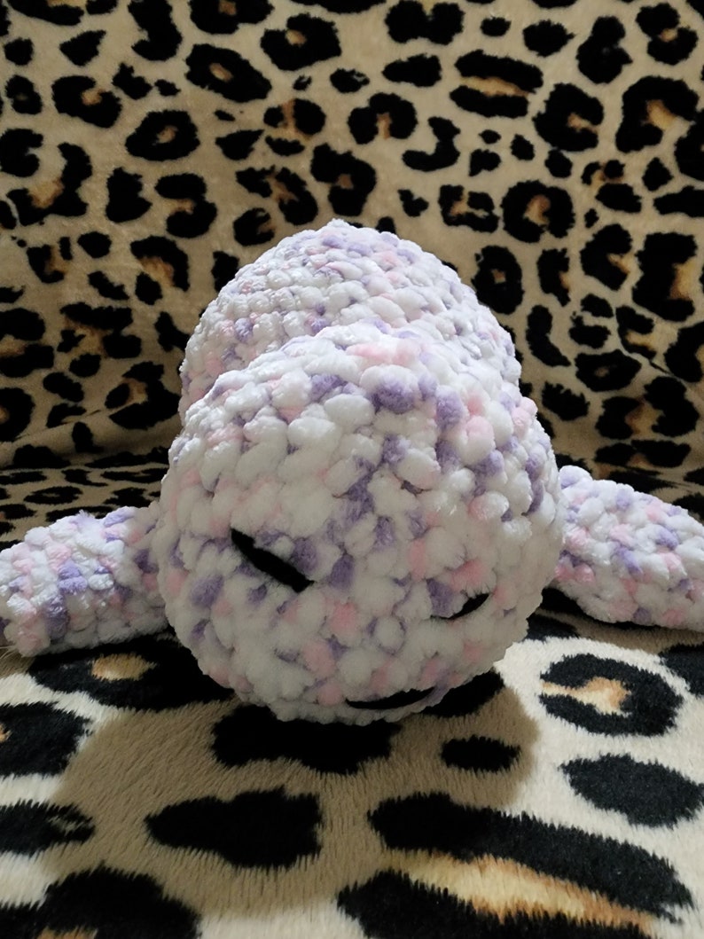 May include: A plush toy shaped like a seal with a white, pink, and purple speckled fur. The toy has black eyes and a closed mouth.