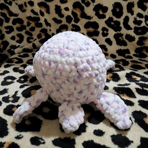 May include: A crocheted plush toy in the shape of a turtle with a white and purple body and four legs. The toy is sitting on a leopard print fabric.