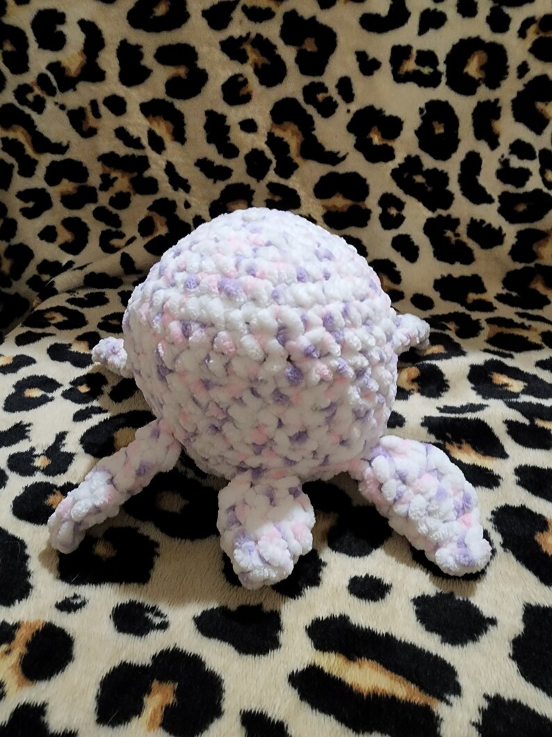 May include: A crocheted plush toy in the shape of a turtle with a white and purple body and four legs. The toy is sitting on a leopard print fabric.