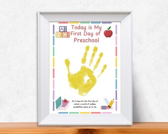 May include: A framed print with the text "Today is My First Day of Preschool." The print features a yellow handprint, an apple, and colorful borders. The bottom of the print has the text "As I step into the first day of school, a world of endless possibilities opens up to me."