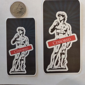 May include: Two rectangular stickers featuring a black and white image of the statue of David, with a red banner across the image that says "Censored". The stickers are on a white surface, with a U.S. quarter for size comparison.
