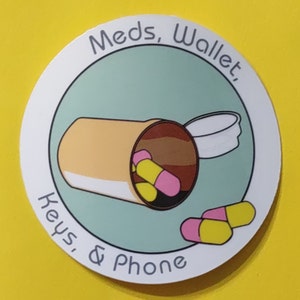 May include: A white round sticker with a light blue circle in the center. The text "Meds, Wallet, Keys, & Phone" is written in a circle around the light blue circle. Inside the light blue circle is a cartoon illustration of a pill bottle with pills spilling out. The pills are yellow and pink.