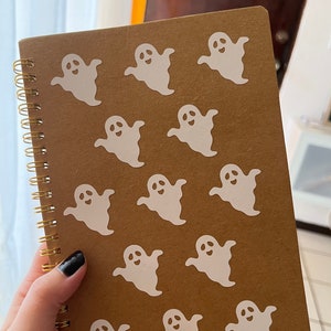 Puede incluir: Un cuaderno marrón con encuadernación en espiral y pegatinas de fantasmas blancas en la portada.