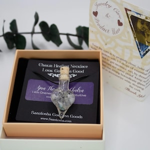 May include: A black cord necklace with a heart-shaped glass bottle pendant filled with crystals and a cork stopper. The pendant is labeled "Yina The Chakra" and "I Am Oneness, Intuitive". The necklace is in a black and white box with the text "Chakra Healing Necklace Look Good, Feel Good" and "Saantosha Goddess Goods www.Saantosha.com".